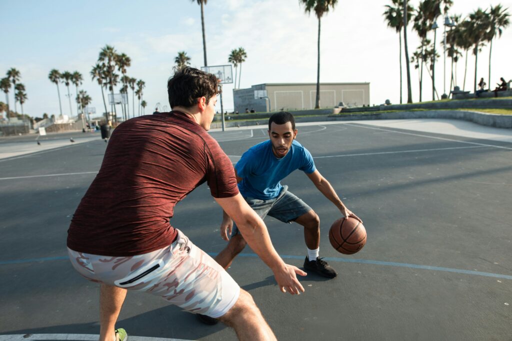 Action shot of two men in a basketball game on an outdoor court with palm trees.