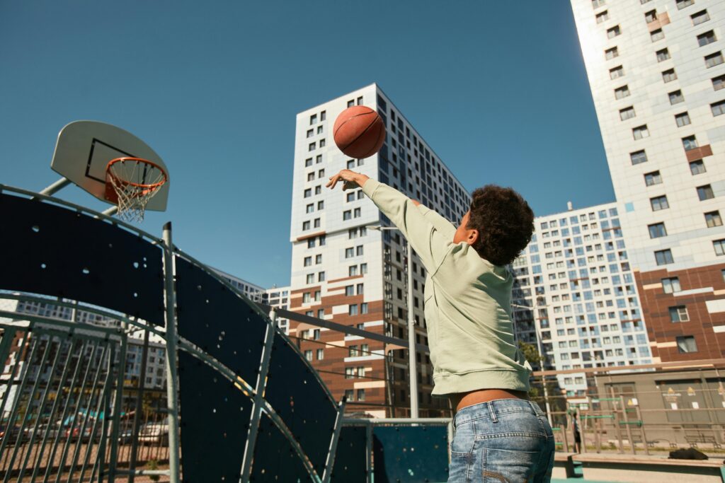 A child playing basketball in a city playground surrounded by tall buildings, enjoying a sunny day.