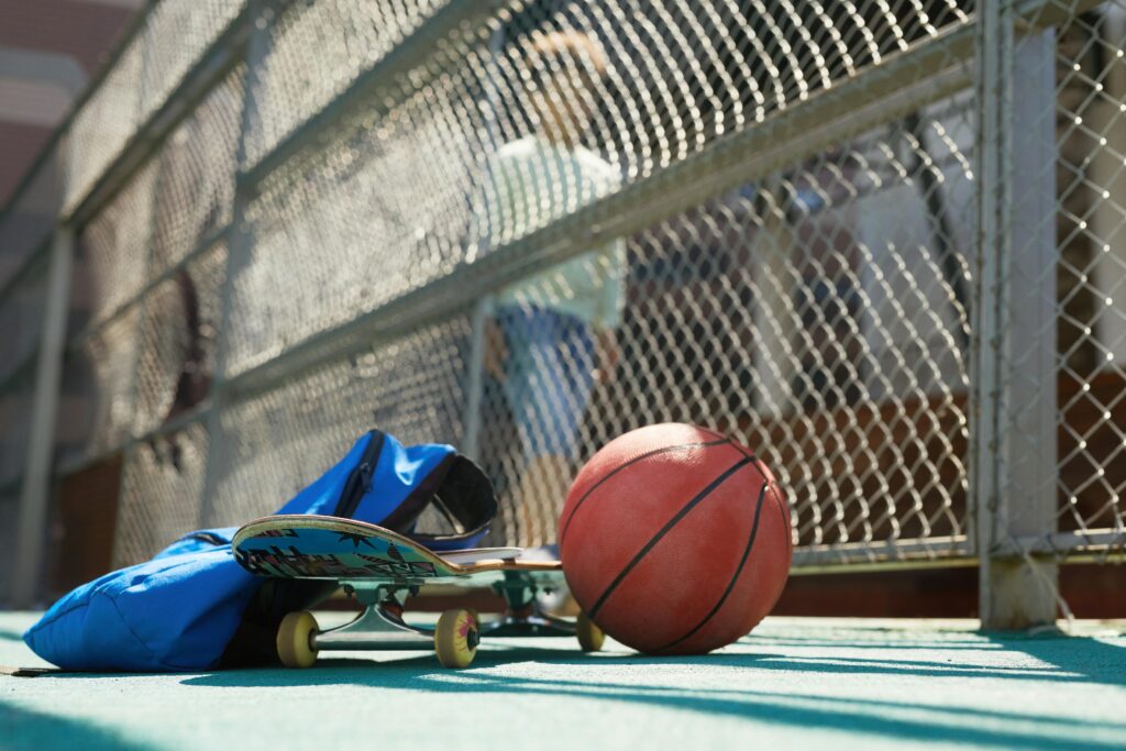 Skateboard, basketball, and blue bag placed near a chain-link fence, capturing active urban lifestyle.