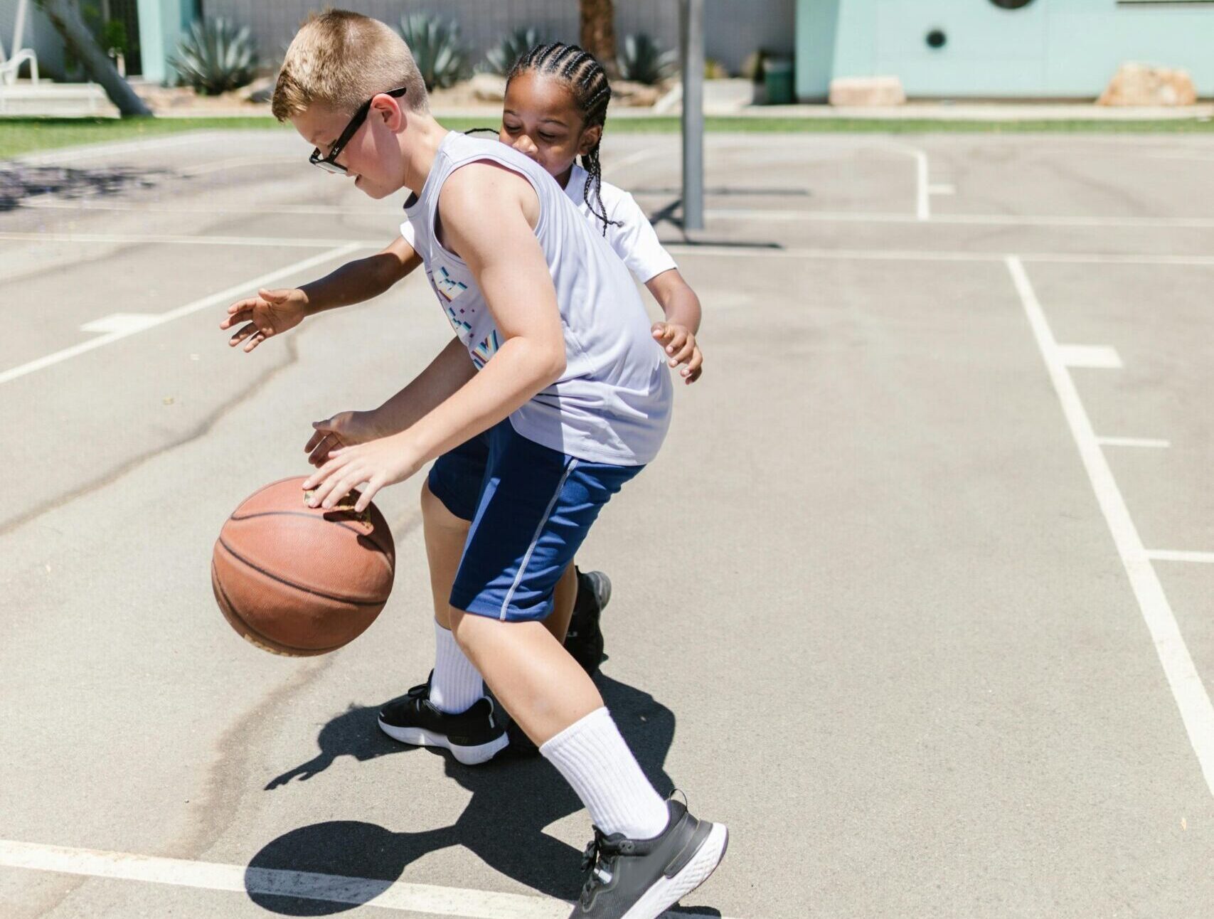 Two kids playing basketball on an outdoor court on a sunny day.