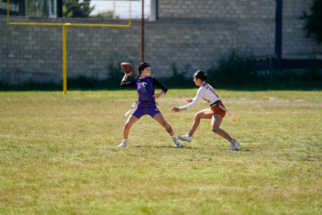 Two women engaged in a competitive outdoors flag football game on a field.